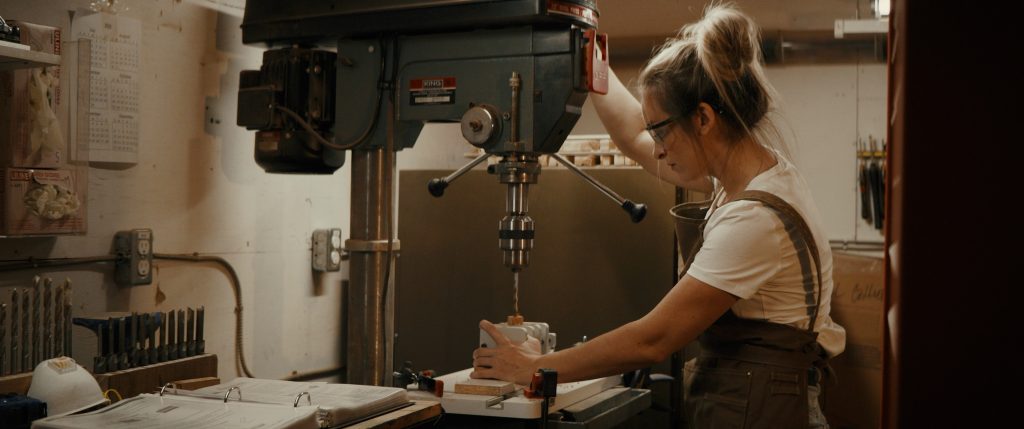 Woman drilling hole into wooden pen blank from William Wood-Write Ltd using a drill press and brad point drill, pictured in dim lighting from a wide angle.
