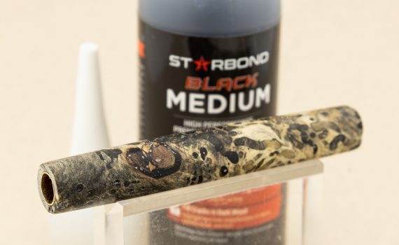 A bottle of black CA glue with a finished burl pen barrel on a display stand