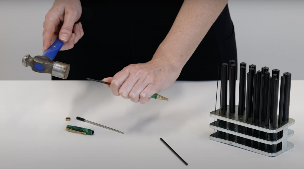 Using a hammer and pen disassembly transfer punch set to tap the gold nib pen kit part out of a gold budget fancy slimline pen kit, pictured next to a pen disassembly transfer punch set and a hammer on a desk.