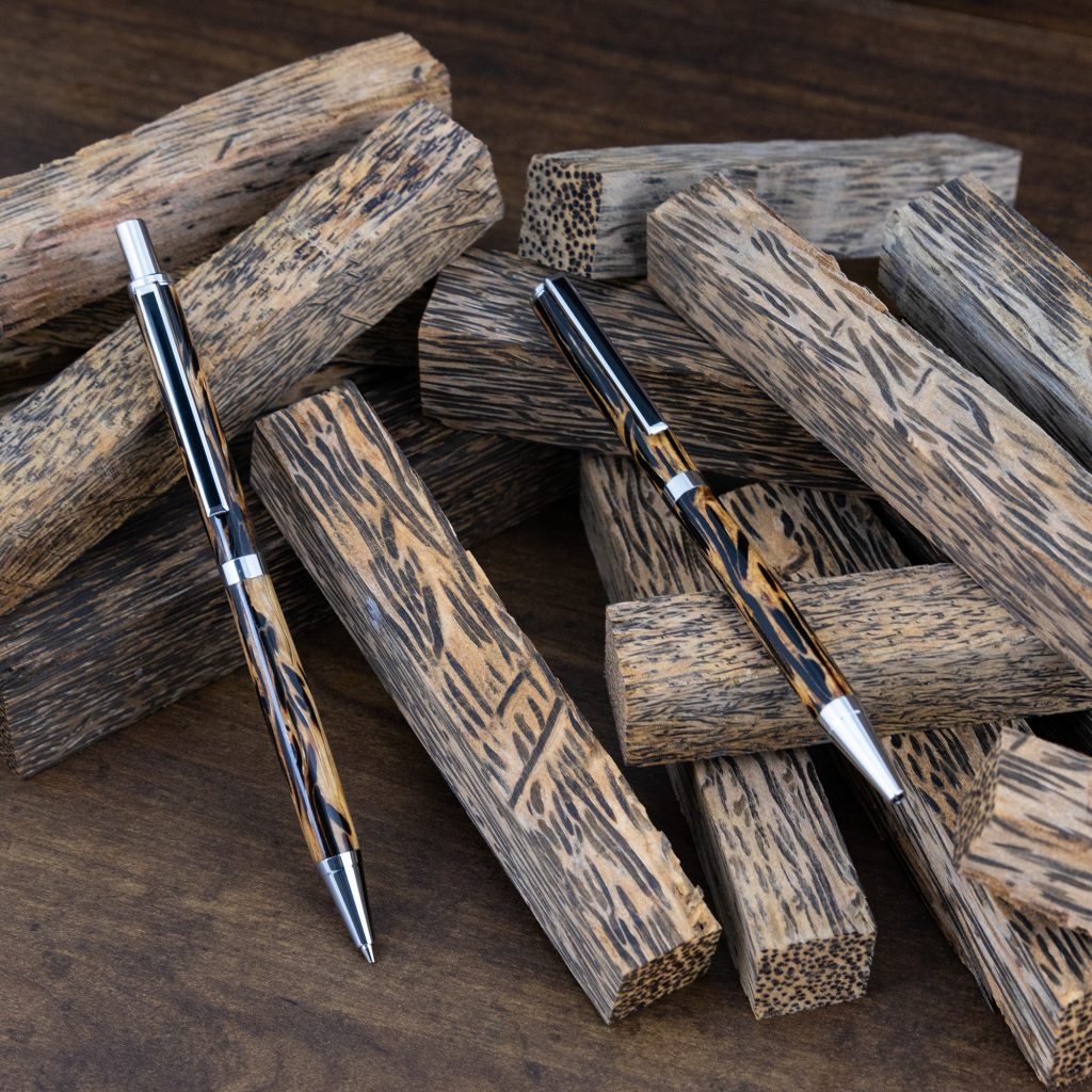 Two handmade pens made of stabilized black palm wood lean on an assortment of black palm blanks.