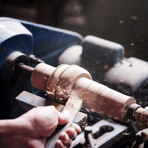 A hand holds a chisel against a spindle on a wood turning lathe, making sawdust fly.