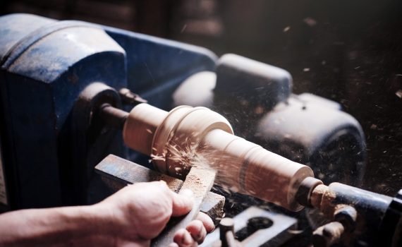 A hand holds a chisel against a spindle on a wood turning lathe, making sawdust fly.