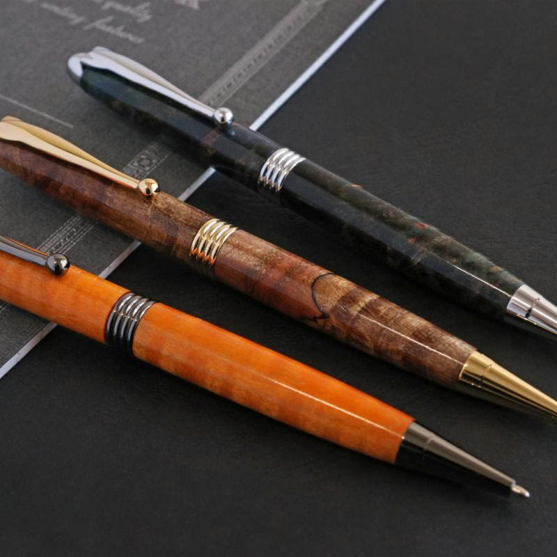 Three slimline pens made with wood on a lathe lying across the edge of a book. One is orange, one variegated brown and one black.