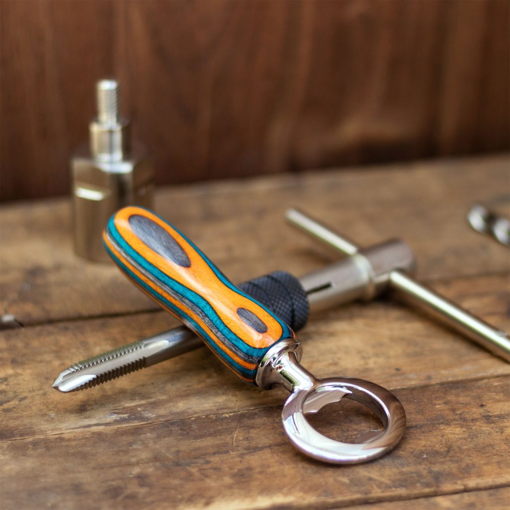 A handmade chrome bottle opener with a wooden handle rests on a drill and tap set with a weathered wood background