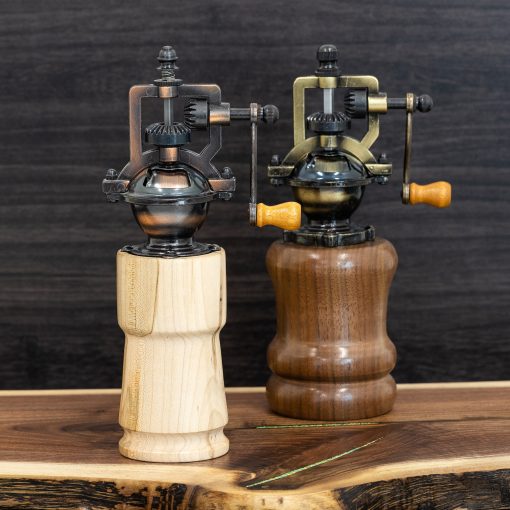 Two pepper grinders, one light wood and one dark wood, are displayed on a wooden background. The tops are fancy crank mechanisms in antique bronze.