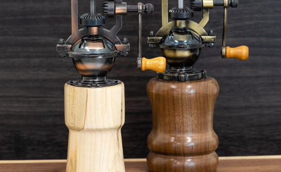 Two pepper grinders, one light wood and one dark wood, are displayed on a wooden background. The tops are fancy crank mechanisms in antique bronze.