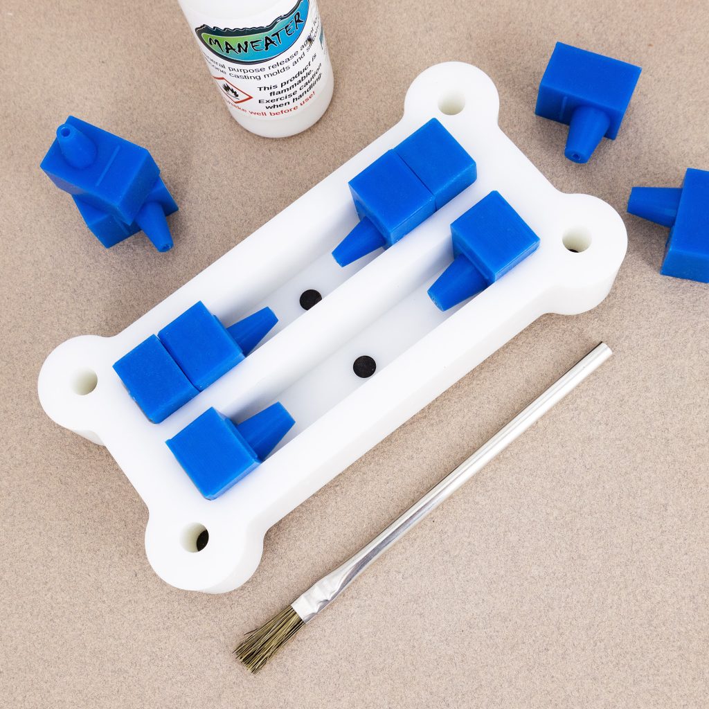 A white plastic mold for making resin pen blanks with two chambers and blue silicone stops and mounts with a paintbrush on the side.