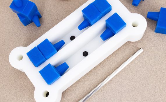 A white plastic mold for making resin pen blanks with two chambers and blue silicone stops and mounts with a paintbrush on the side.