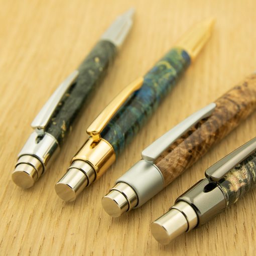 Four wood turned custom pens in various burl are displayed on a yellow wooden background. The push buttons are chrome and gold.