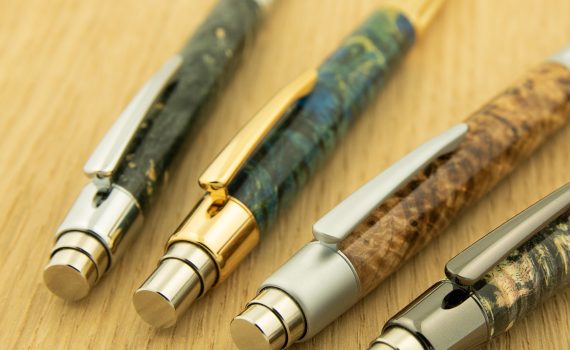 Four wood turned custom pens in various burl are displayed on a yellow wooden background. The push buttons are chrome and gold.