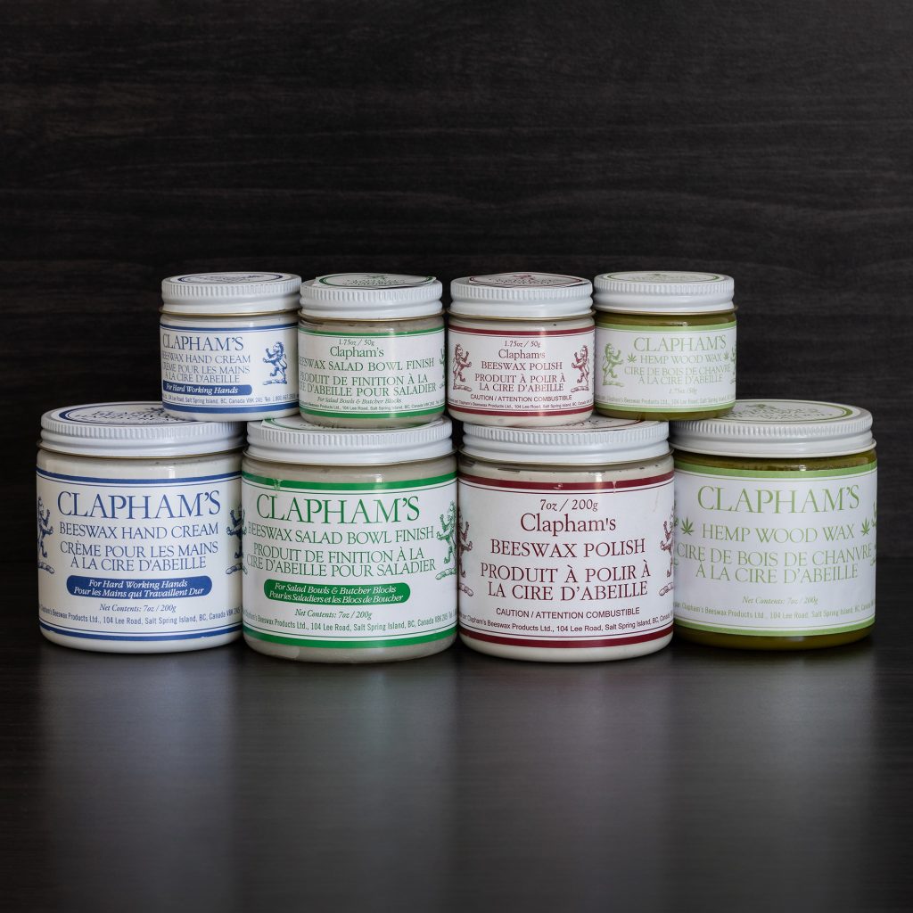 Large and small glass jars with white lids stacked on a black background. The labels on the jars say Claphams wood wax.