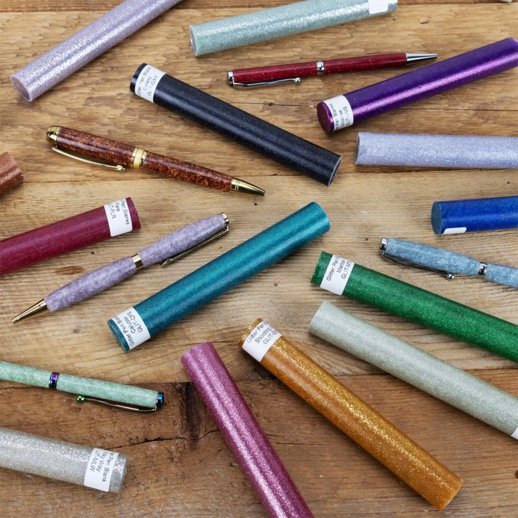 Glittery pen blanks in many colours scattered on a wooden surface with several glitter handmade custom pens