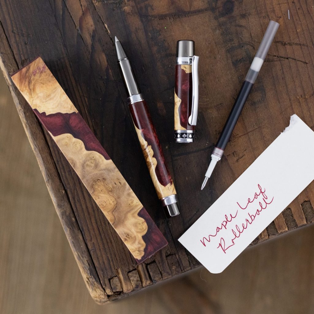 A maroon and burl pen blank, handmade wood turned pen and ink insert wit on a rough wooden surface alongside a piece of paper that says "Maple Leaf Rollerball" in maroon ink.