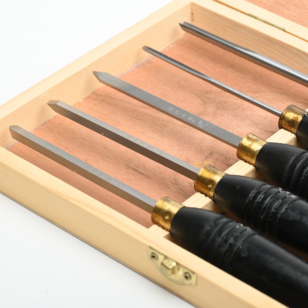 A set of 5 steel chisels with black wooden handles sit in a wooden display box