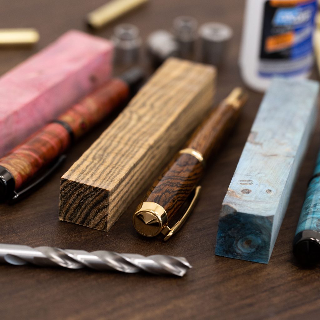 A selection of woodturning materials on a brown table, including two custom made pens, three exotic wood pen blanks, a drill bit and CA glue.