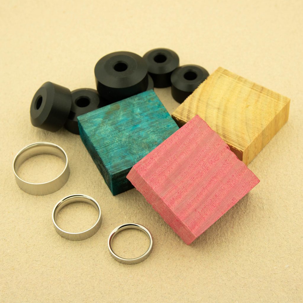 Three steel rings are displayed beside three stabilized wood blanks in yellow, blue and red, and a set of bushings in the background.