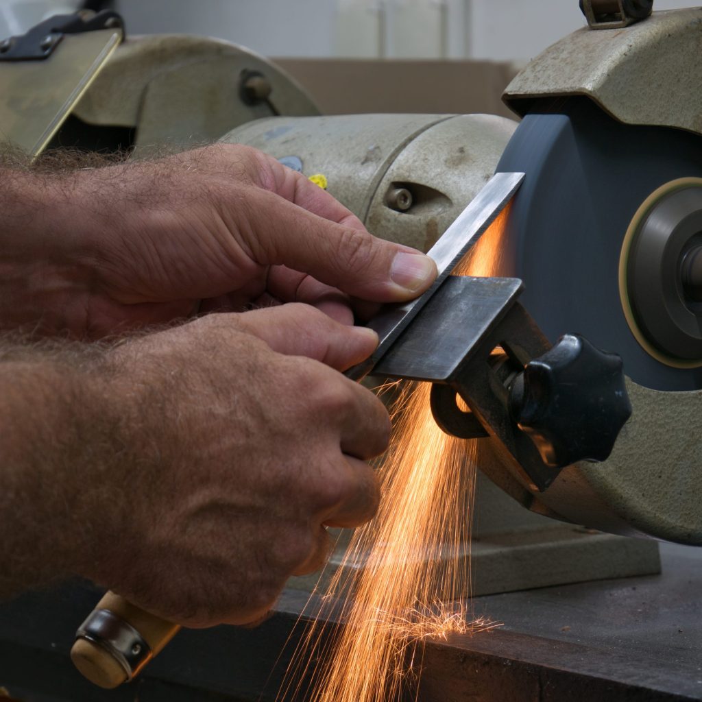 A steel chisel is held with two hands against the wheel of a bench grinder with sparks flying off of it.