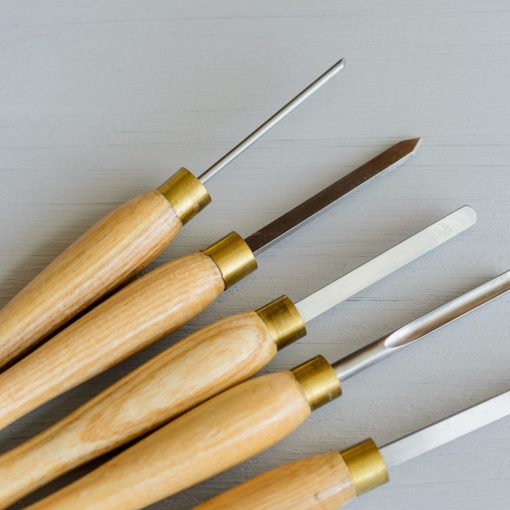 Five woodturning chisels with steel tips and yellow wooden handles on a grey background.