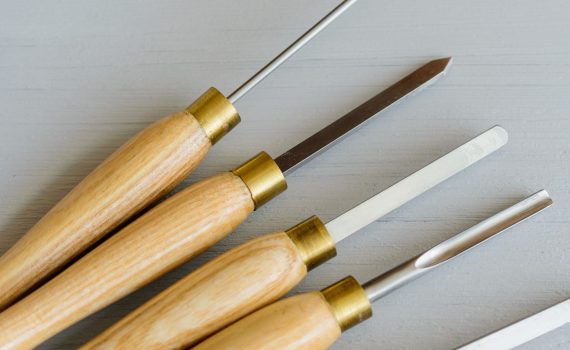 Five woodturning chisels with steel tips and yellow wooden handles on a grey background.