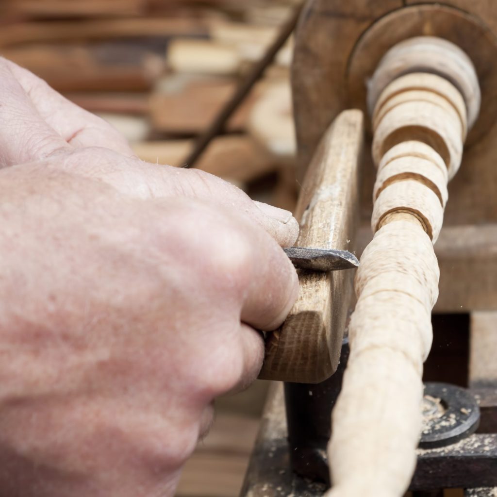 A closeup of hand holding a chisel up to a wooden spindle on a running wood lathe.