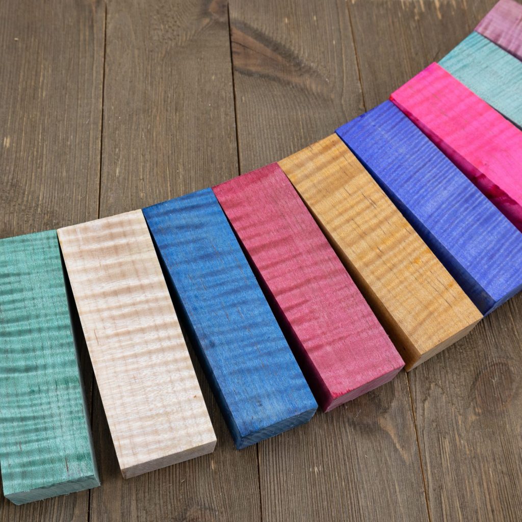 Curly maple blocks stabilized and dyed various colours lined up on a wooden background.