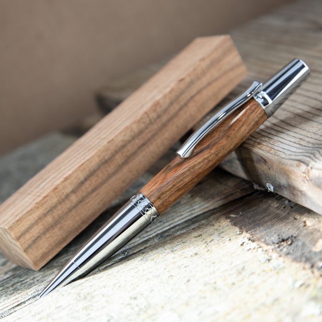 A handmade wood pen made from rosewood beside a rosewood pen blank resting on a wooden surface.