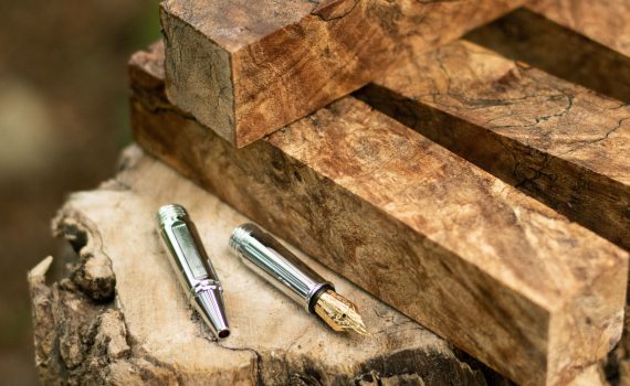 Spalted Maple pen blanks are piled on a tree stump next to a fountain pen nib and roller ball nib.